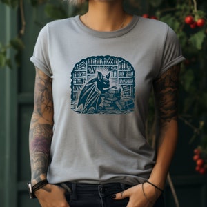 May include: A gray t-shirt with a blue graphic of a bat reading a stack of books in a library.