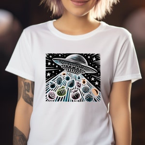 Puede incluir: Camiseta blanca con un gráfico en blanco y negro de un OVNI con un fondo estrellado. El OVNI está proyectando una variedad de frutas, incluyendo manzanas, peras y bayas.