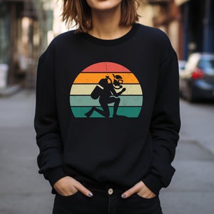 Peut inclure: Un sweat-shirt noir avec un graphique vintage d'un plongeur devant un coucher de soleil. Le graphique est dans des tons d'orange, jaune, vert et bleu.