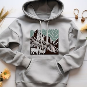 May include: A gray hooded sweatshirt with a graphic print of a mountain landscape and llamas. The print features a brown mountain with a waterfall, green hills, and a brown path. Three white llamas are walking on the path.