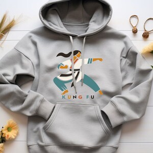 Peut inclure: Un sweat à capuche gris avec un motif représentant une femme en position de combat d'arts martiaux. Le texte "KUNG FU" est imprimé sous le motif.