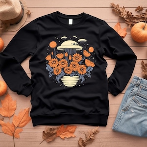 Puede incluir: Sudadera negra con un gráfico de un platillo volante flotando sobre una maceta de flores naranjas. Las flores están rodeadas de estrellas y el platillo tiene un rayo de luz que sale de él.
