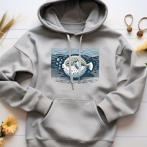 Puede incluir: Una sudadera con capucha gris con un gráfico de un pez globo nadando en el océano. El gráfico está en tonos de azul, blanco y negro.