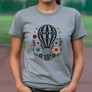 Peut inclure: Un t-shirt gris avec une illustration noire et blanche d'une montgolfière entourée de fleurs. Les fleurs sont dans des tons de bleu, rose et orange.