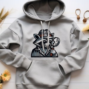Puede incluir: Una sudadera con capucha gris con un estampado gráfico de un mapache con un sombrero y sosteniendo una lupa. El mapache está mirando a través de la lupa.