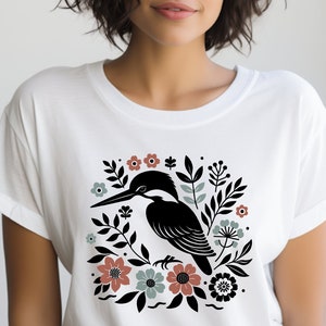 May include: White t-shirt with a black silhouette of a bird surrounded by flowers and leaves. The bird is perched on a branch and has a long beak. The flowers are in shades of pink, red, and blue.