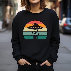 May include: A black sweatshirt with a retro-style graphic of a UFO and an alien in front of a sunset. The graphic has a vintage, distressed look.