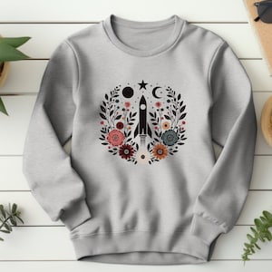 May include: A gray sweatshirt with a black rocket ship surrounded by a floral wreath. The wreath includes black, white, red, and orange flowers. The rocket ship is launching into a black sky with a white star and a crescent moon.