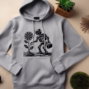 May include: A gray hoodie with a black robot watering a sunflower. The robot is holding a bucket in one hand and a watering can in the other. The design is printed on the front of the hoodie.