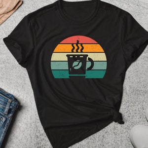 Puede incluir: Camiseta negra con un gráfico de puesta de sol retro que presenta una taza de café con vapor saliendo de ella.