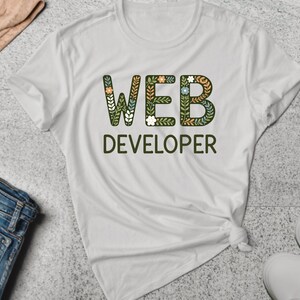May include: White t-shirt with the text "WEB DEVELOPER" in green letters. The letters are decorated with floral patterns in shades of green, orange, and white.