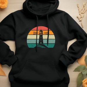 May include: Black hooded sweatshirt with a vintage-style graphic of a lighthouse in front of a sunset. The graphic has a faded, distressed look with orange, yellow, blue, and green colors.