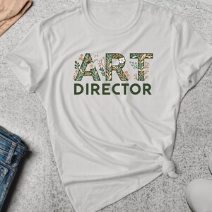 以下が含まれることがあります： 前面に「ART DIRECTOR」と綴られた緑と茶色の花柄の白いTシャツです。