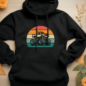 Puede incluir: Sudadera con capucha negra con un gráfico retro de un monster truck. El diseño muestra un camión blanco con ruedas grandes, sobre un fondo de puesta de sol en un diseño circular con rayas naranjas, amarillas y turquesas.