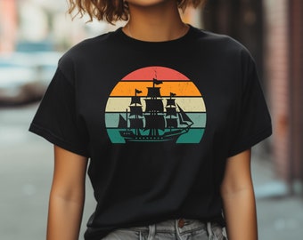 Puede incluir: Camiseta negra con un gráfico de estilo vintage de un velero frente a una puesta de sol. La puesta de sol es un degradado multicolor de naranja, amarillo y azul.