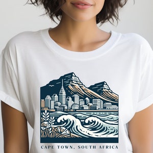 May include: White t-shirt with a graphic design of the Cape Town skyline, featuring Table Mountain, buildings, and ocean waves. The text "CAPE TOWN, SOUTH AFRICA" is printed below the graphic.