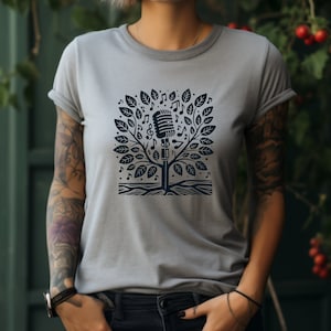 Puede incluir: Una camiseta gris con un gráfico en blanco y negro de un micrófono dentro de un árbol con notas musicales. El árbol tiene raíces y hojas.