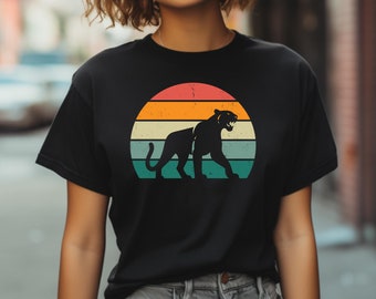 Puede incluir: Camiseta negra con un gráfico de estilo vintage de una silueta de pantera negra frente a una puesta de sol. La puesta de sol es un semicírculo con rayas naranja, amarillo y azul.