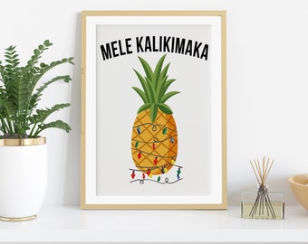 Op de afbeelding: Een ingelijste print met een gele ananas versierd met kleurrijke kerstverlichting. De tekst "MELE KALIKIMAKA" staat in het zwart boven de ananas. De print staat op een witte plank met een potplant en een diffuser.