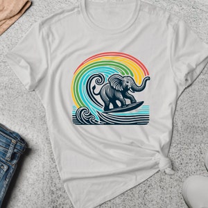 Puede incluir: Camiseta blanca con un gráfico de un elefante surfeando en una ola con un arcoíris en el fondo.