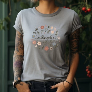 Puede incluir: Una camiseta azul claro con un diseño floral y el texto "calígrafo" en letra cursiva.