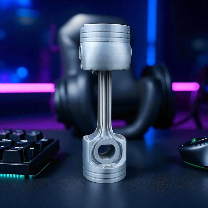 A silver-colored piston-shaped stand with a plain top, no skull, stands upright on a dark desk with a glowing keyboard and mouse.