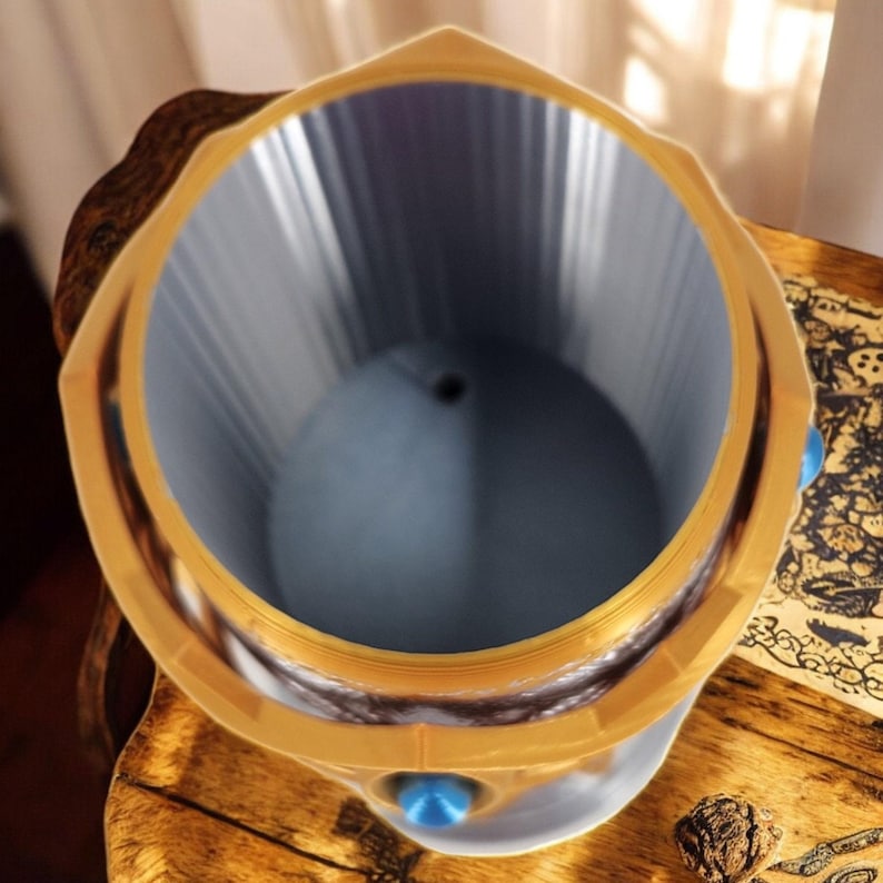 An overhead view of a gold and silver 3D-printed goblet with a blue interior, sitting on a wooden table next to an open book.