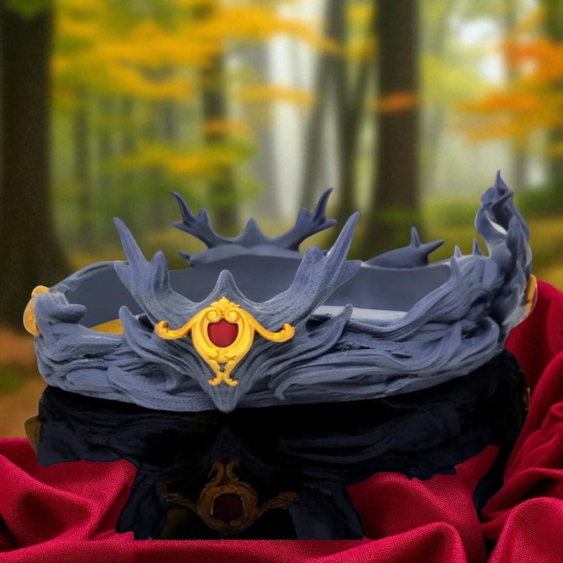 A dark gray, textured crown with a red and gold central emblem rests on a reflective red fabric, with a blurred forest background.