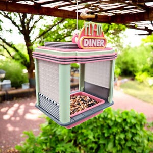 A pink, mint green, and gray retro diner-style bird feeder with "FLY-IN DINER" on its sign hangs from a wooden beam, filled with birdseed, with green foliage in the background.