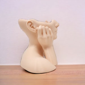 Op de afbeelding: Een beige keramische sculptuur van een hoofd en schouders van een persoon met de handen op de kin. De sculptuur is ontworpen om een plantenbak te zijn, met een opening in de bovenkant van het hoofd.