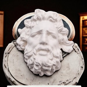 May include: A white bust of a bearded man with curly hair and a serene expression. The sculpture features a detailed beard and hair, set against a circular backdrop with a dark blue and gold trim. The bust is mounted on a white, weathered base.