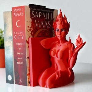 May include: A red, flame-shaped bookend featuring a stylized female figure with fiery hair and holding a flame. The bookend is positioned next to several books, including one with the title "Crescent City" and the author's name "Sarah J. Maas."