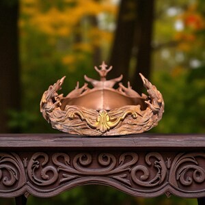 A brown, textured crown with a gold-colored central emblem is displayed on a dark carved wooden table, with a blurred autumn-colored background.