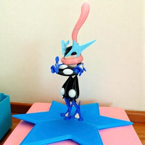 May include: A blue, black, and white figurine of a character with a long pink tongue, wearing a blue helmet and a black cloak. The figurine is standing on a blue star-shaped base, set against a pink surface.