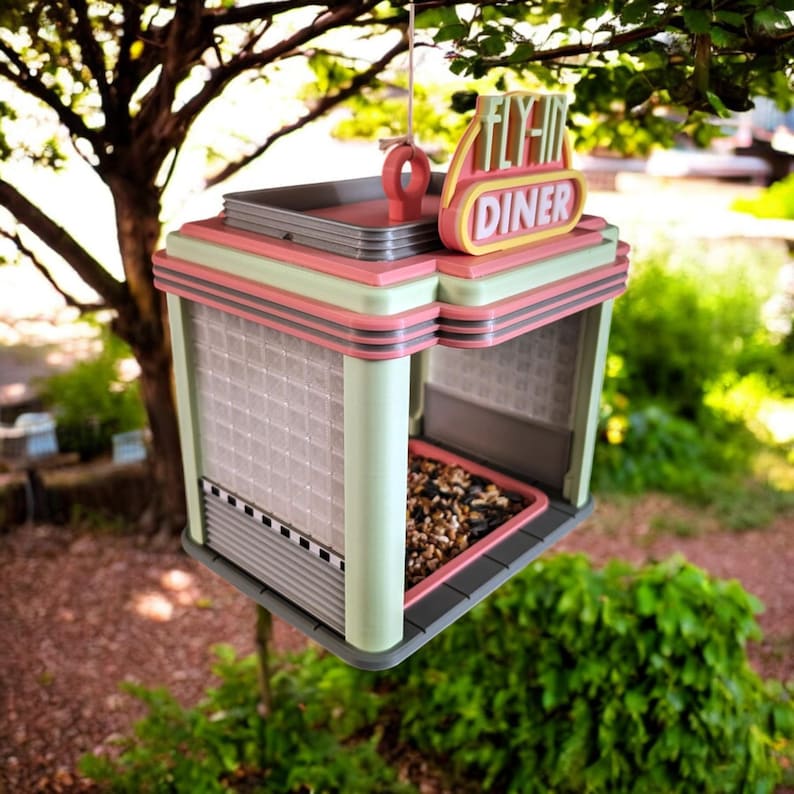 A pink, mint green, and gray retro diner-style bird feeder with "FLY-IN DINER" on its sign hangs from a tree branch, filled with birdseed.