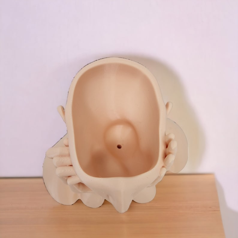 Op de afbeelding: Een beige keramische plantenpot in de vorm van een mensenhoofd met handen die het gezicht bedekken. De pot heeft een gat bovenaan voor drainage.