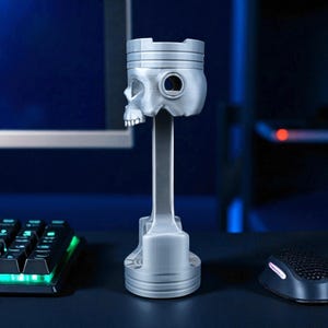 A silver-colored piston-shaped headphone stand with a skull head is shown from the side on a dark desk, with a glowing keyboard and mouse.