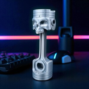 A silver-colored piston-shaped headphone stand with a skull head is angled on a dark desk, with a glowing keyboard and mouse in the background.