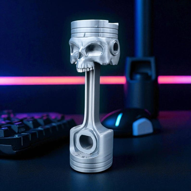 A silver-colored piston-shaped headphone stand with a skull head is angled on a dark desk, with a glowing keyboard and mouse in the background.