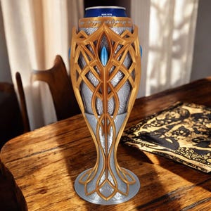 A gold and silver 3D-printed goblet, holding a blue soda can, sits on a wooden table next to an open book with intricate patterns.