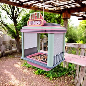 A pink, mint green, and gray retro diner-style bird feeder with "FLY-IN DINER" on its sign hangs from a wooden beam, filled with birdseed.