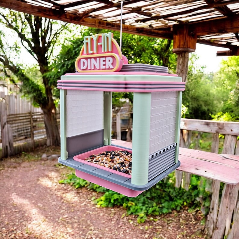 A pink, mint green, and gray retro diner-style bird feeder with "FLY-IN DINER" on its sign hangs from a wooden beam, filled with birdseed.