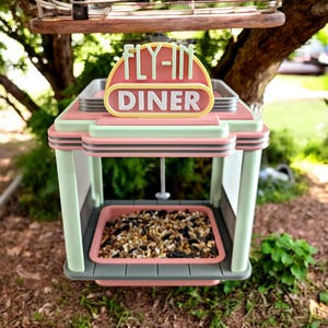 A pink, mint green, and gray retro diner-style bird feeder with "FLY-IN DINER" on its sign sits on the ground near a tree, filled with birdseed.