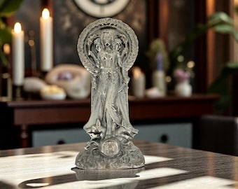 May include: A silver-toned statue depicting a three-headed figure standing within a circular frame. The figure wears long, flowing robes and holds objects in its hands. The base of the statue is textured and features a circular emblem at the center.