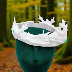 A white, textured crown with a green gem is shown from the side on a dark green velvet head form, with a blurred green forest in the background.