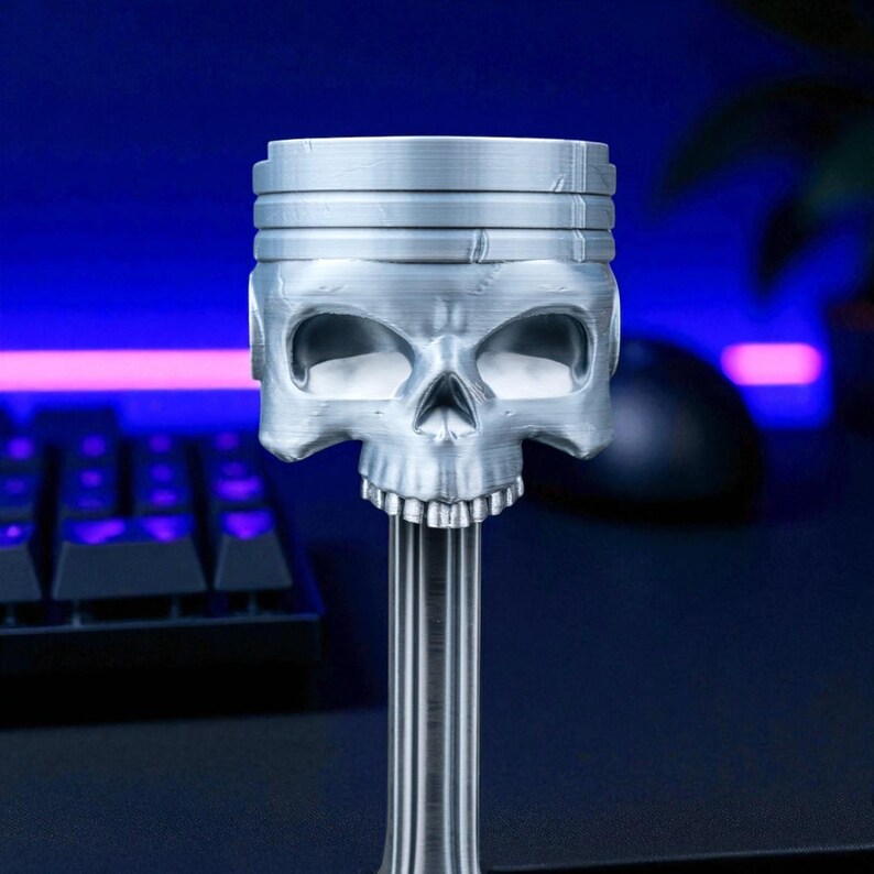 A close-up of the silver-colored skull head of the piston-shaped headphone stand, showing textured details, on a dark desk.