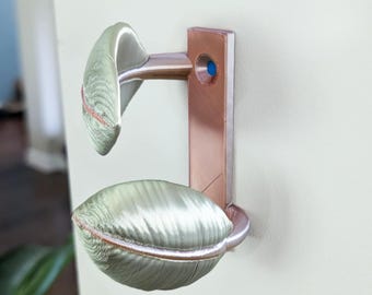 May include: A decorative wall-mounted object with a copper-colored metal frame and two light green, oval-shaped fabric elements. The fabric has a textured appearance with a contrasting trim. A small blue accent is visible.