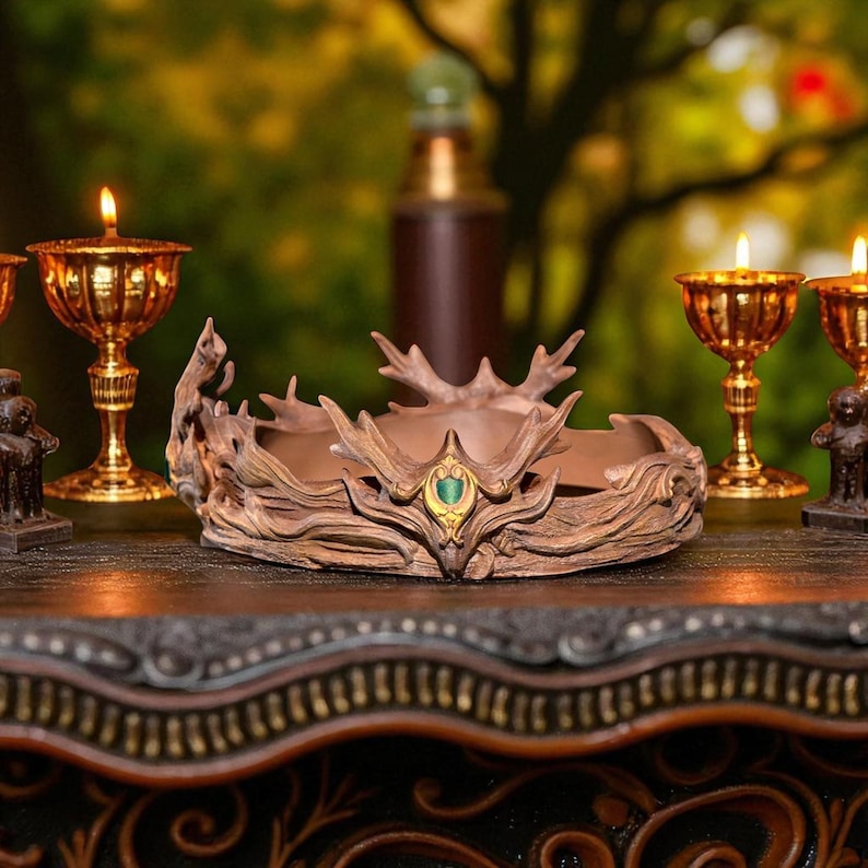 A brown, textured crown with a green gem in the center sits on a dark wooden surface, flanked by two golden chalices with lit candles.