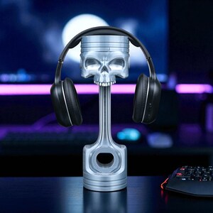 A silver-colored piston-shaped headphone stand with a skull head holds black wireless headphones, with a glowing keyboard and mouse visible on a dark desk.