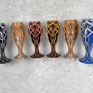 Six 3D-printed goblets in various colors, including silver, gold, copper, and blue, with intricate elven designs, are arranged in a row on a gray textured surface.